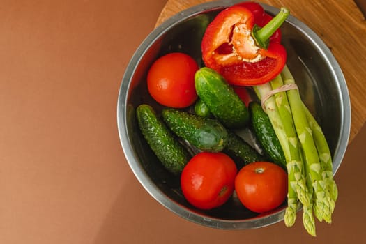 Vegetables lie in a metal bowl: tomatoes, asparagus, cucumbers, red bell peppers. on a wooden board and brown background. place for text. view from above.