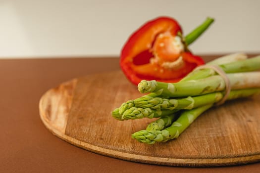 branches of fresh green asparagus on a wooden board, half of a red big pepper in the background. brown and light beige background. Basic trend concept with copy space