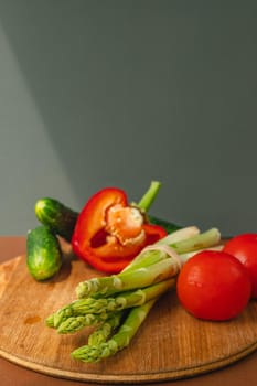 Vegetables lie on a wooden board: tomatoes, asparagus, cucumbers, red bell peppers. brown, dark gray background. place for text