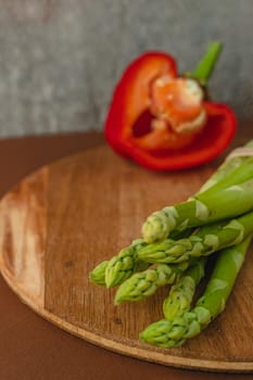 branches of fresh green asparagus on a wooden board, red big pepper on the background. brown background, top view. Basic trend concept with copy space