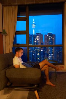Guy rest in cozy sofa with laptop against window with stunning city landscape. Kuala Lumpur, Malaysia - 04.01.2020