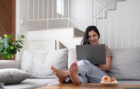 Young business freelance Asian woman working on laptop checking social media while lying on the sofa when relax in living room at home..