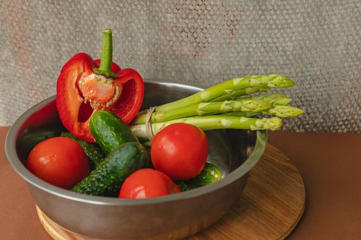 Vegetables lie in a metal bowl: tomatoes, asparagus, cucumbers, red bell peppers . on a wooden board and brown background. back gray background. place for text.