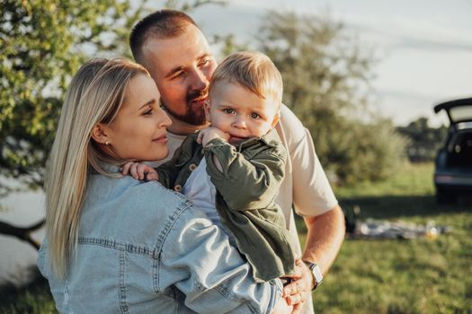 Portrait of Young Family Outdoors, Father and Mother Hugging Their Toddler Son that Sadly Holding His Finger in the Mouth