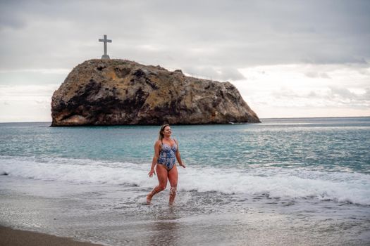 A plump woman in a bathing suit enters the water during the surf. Alone on the beach, Gray sky in the clouds, swimming in winter