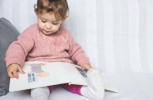 Little baby girl reading a picture book on a white sofa