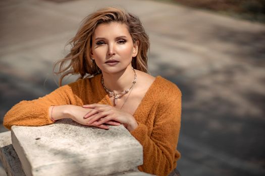 A middle-aged woman looks like a good blonde with curly beautiful hair and makeup on the background of the building. She is wearing a yellow sweater