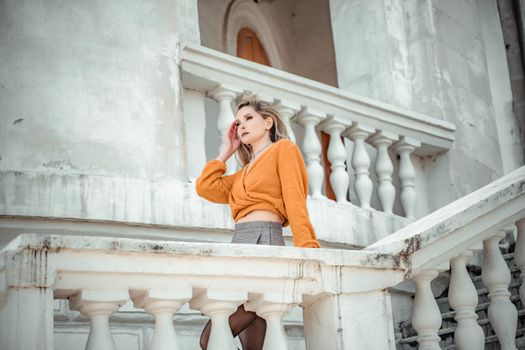 A middle-aged woman looks like a good blonde with curly beautiful hair and makeup on the background of the building. She is wearing a yellow sweater