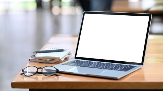 Computer laptop with white empty display, eyeglasses and notebooks on wooden office desk.