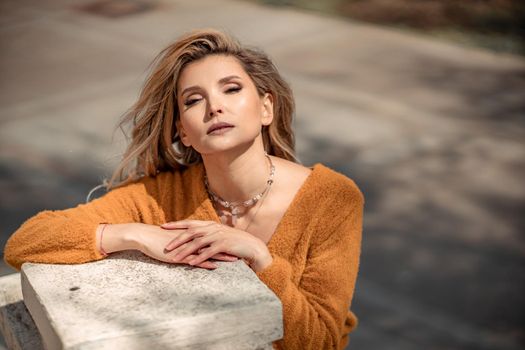 A middle-aged woman looks like a good blonde with curly beautiful hair and makeup on the background of the building. She is wearing a yellow sweater