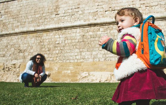 Toddler wearing a backpack on the grass with her mother in the background