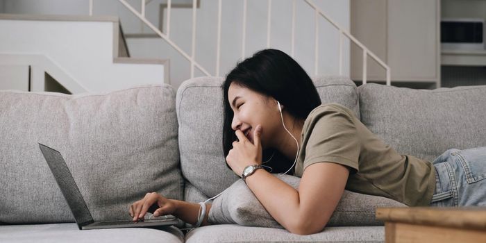 Asian woman lying on the sofa in the living room listening to music on a laptop device woman relaxing on the sofa at home View messages on the computer screen on the device. communication concept.