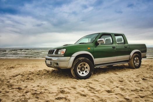Green pick-up truck parked on the sandy beach with sea, blue sky and clouds in the background