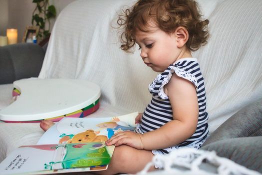 Little baby girl reading a picture book on a white sofa