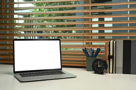 Laptop computer with blank screen, alarm clock, pencil holder and book on white table.