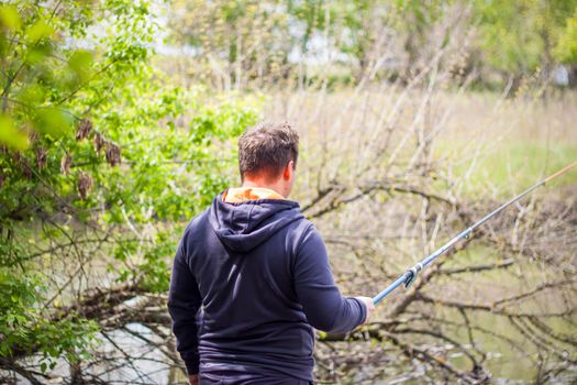 A male fisherman stands by the river with a simple fishing rod, a man is resting while fishing, the spring sun is bright. fishing