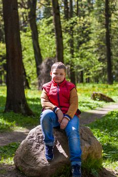 A young man in a sleeveless red jacket is sitting on a huge stone in a pine forest in spring. The magical light from the sun's rays falls behind the boy. Spring