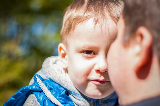 The child is in the father's arms, hiding behind his back. Interaction. Facial expression. Take a walk in the green park in the fresh air. The magical light from the sun's rays is left behind. Spring