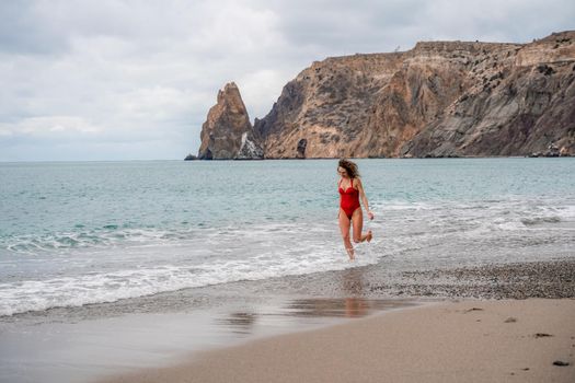 A beautiful and sexy brunette in a red swimsuit on a pebble beach, Running along the shore in the foam of the waves.