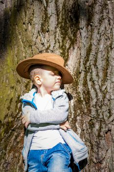 Cute boy posing in a cowboy hat in the woods by a tree. The sun's rays envelop the space. Interaction history for the book. Space for copying. Selective focus.