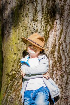 Cute boy posing in a cowboy hat in the woods by a tree. The sun's rays envelop the space. Interaction history for the book. Space for copying. Selective focus.