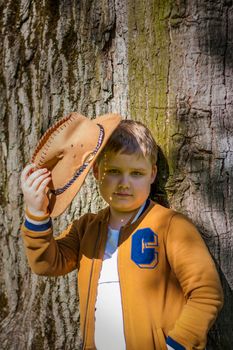 Cute boy posing in a cowboy hat in the woods by a tree. The sun's rays envelop the space. Interaction history for the book. Space for copying. Selective focus.