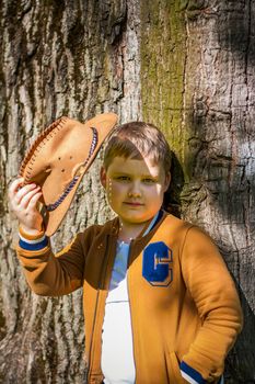 Cute boy posing in a cowboy hat in the woods by a tree. The sun's rays envelop the space. Interaction history for the book. Space for copying. Selective focus.