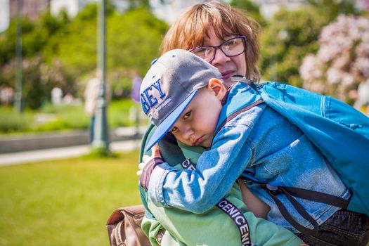A moody child cuddles up to his young grandmother in the park. The woman feels sorry for the boy in her arms. Interactions. Selective focus. Spring
