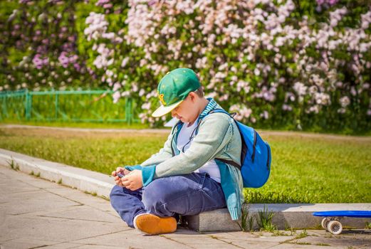 A young man is sitting on the side of a lilac alley and looking at his phone.  Against the background of lilac bushes. Interactions. Selective focus. Spring