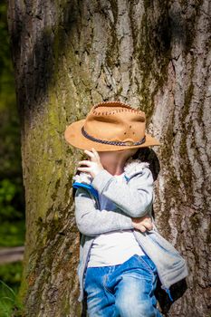Cute boy posing in a cowboy hat in the woods by a tree. The sun's rays envelop the space. Interaction history for the book. Space for copying. Selective focus.