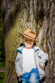 Cute boy posing in a cowboy hat in the woods by a tree. The sun's rays envelop the space. Interaction history for the book. Space for copying. Selective focus.