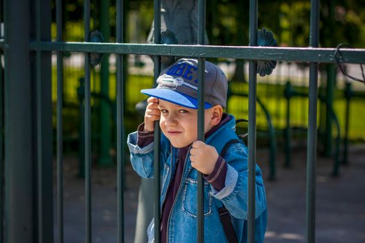 A boy in a baseball cap looks through the iron bars of the fence.  Interactions. Selective focus. Spring