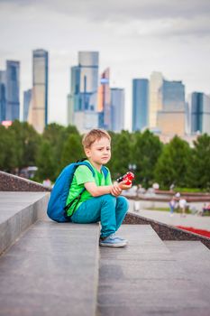 A boy is playing with a toy, sitting on the steps in the open air against the backdrop of skyscrapers and high-rise buildings. Journey. Lifestyle in the city. Center, streets.