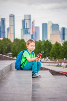 A boy is playing with a toy, sitting on the steps in the open air against the backdrop of skyscrapers and high-rise buildings. Journey. Lifestyle in the city. Center, streets.