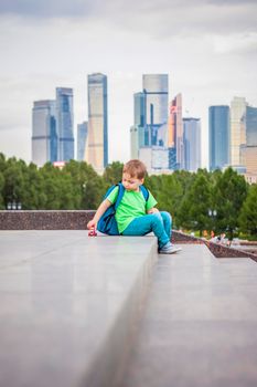 A boy is playing with a toy, sitting on the steps in the open air against the backdrop of skyscrapers and high-rise buildings. Journey. Lifestyle in the city. Center, streets.