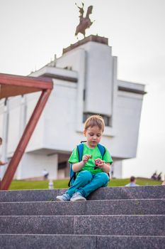 A boy is playing with a toy, sitting on the steps in the open air against the backdrop of skyscrapers and high-rise buildings. Journey. Lifestyle in the city. Center, streets.