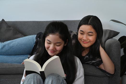 Two happy asian girls sitting in living room and reading book together.
