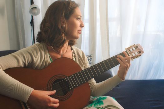 Attractive young woman sitting on a sofa at home playing the Spanish guitar
