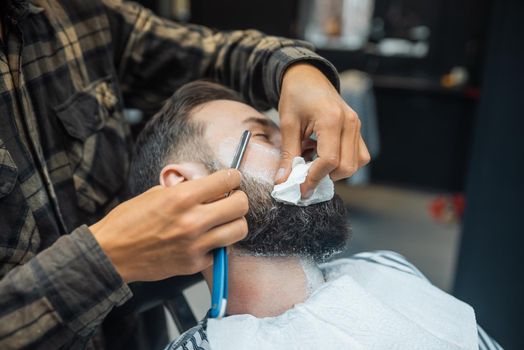 Young bearded man getting shaved with straight edge razor by hairdresser at barbershop