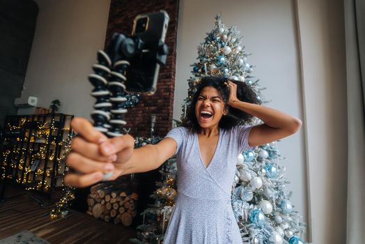 Beautiful Young Smiling Woman near Christmas Tree at the Background Making Video Message or Selfie Concept of Holidays and New Year in Christmas decoration room.