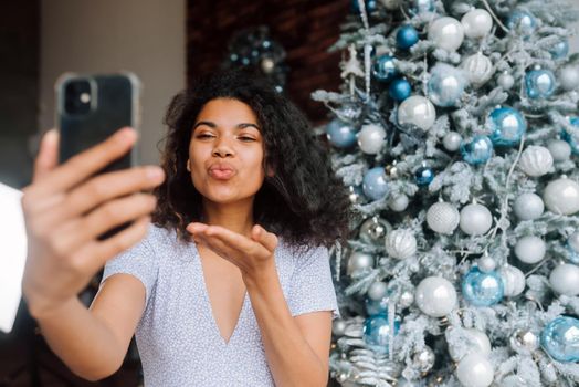 Beautiful Young Smiling Woman near Christmas Tree at the Background Making Video Message or Selfie Concept of Holidays and New Year in Christmas decoration room.