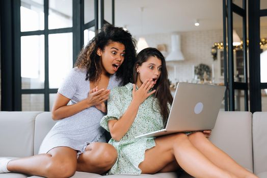 Two beautiful young women relaxing on the living room floor looking at a laptop computer and laughing at something on the screen