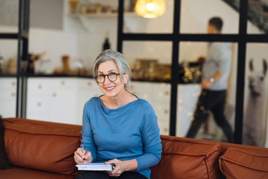 Happy senior woman in glasses writing to notebook or diary at home