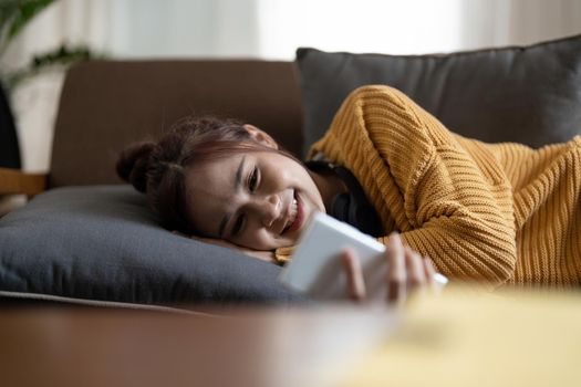 Happy young asian woman lying on sofa at living room and using mobile phone at home.