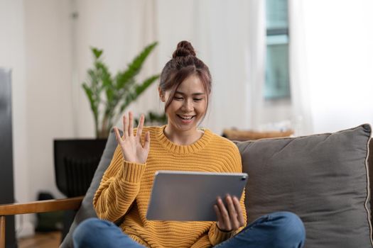 Happy smiling asian young woman in yellow sweater with tablet pc computer having video call and waving hand at home.