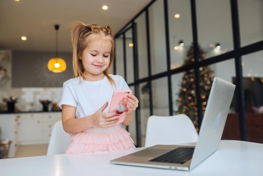 Little kid sit at desk with laptop hold mobile phone at home