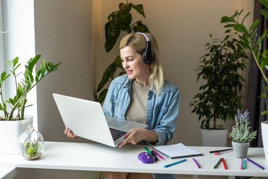 Image of young pleased happy cheerful cute beautiful business woman sit indoors in office using laptop computer listening music with earphones.