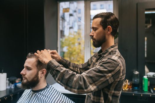 Great time at barbershop. Cheerful young bearded man getting haircut by hairdresser while sitting in chair at barbershop