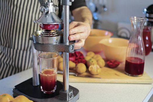 glass of freshly squeezed pomegranate juice, chopped pomegranates, tangerines and a pomegranate juicer at home. Male hands close-up in the process of squeezing juice from fresh fruits.