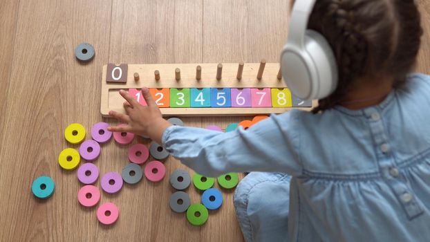 Happy Little Preschool Toothless Girl Playing With Colored Wooden Toy. Kids Learn To Count By Playing Teaches Numbers At Home. Child Listening To Music In Big White Headphones. Childhood, Education.